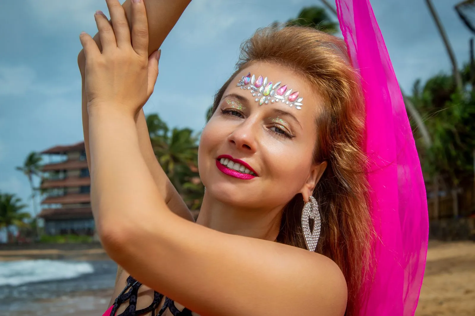 Radiant woman with vibrant makeup and pink scarf enjoying a sunny day on the beach.
